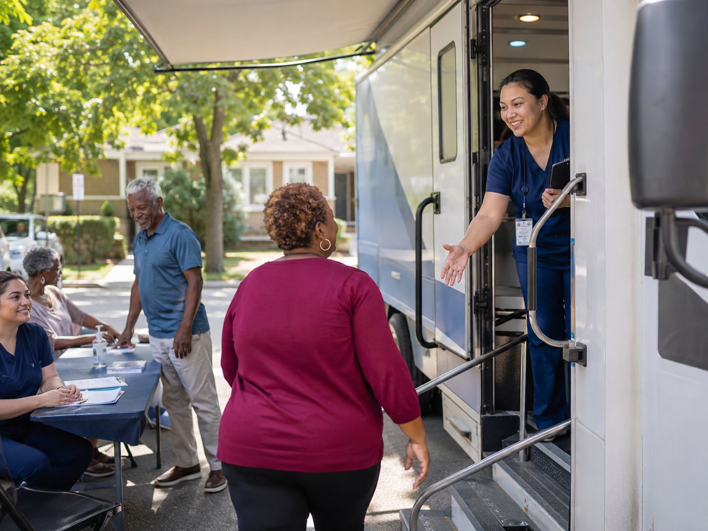 A nurse reaches out to greet a woman approaching a mobile clinic van, with an older man and woman seated at a table nearby.
