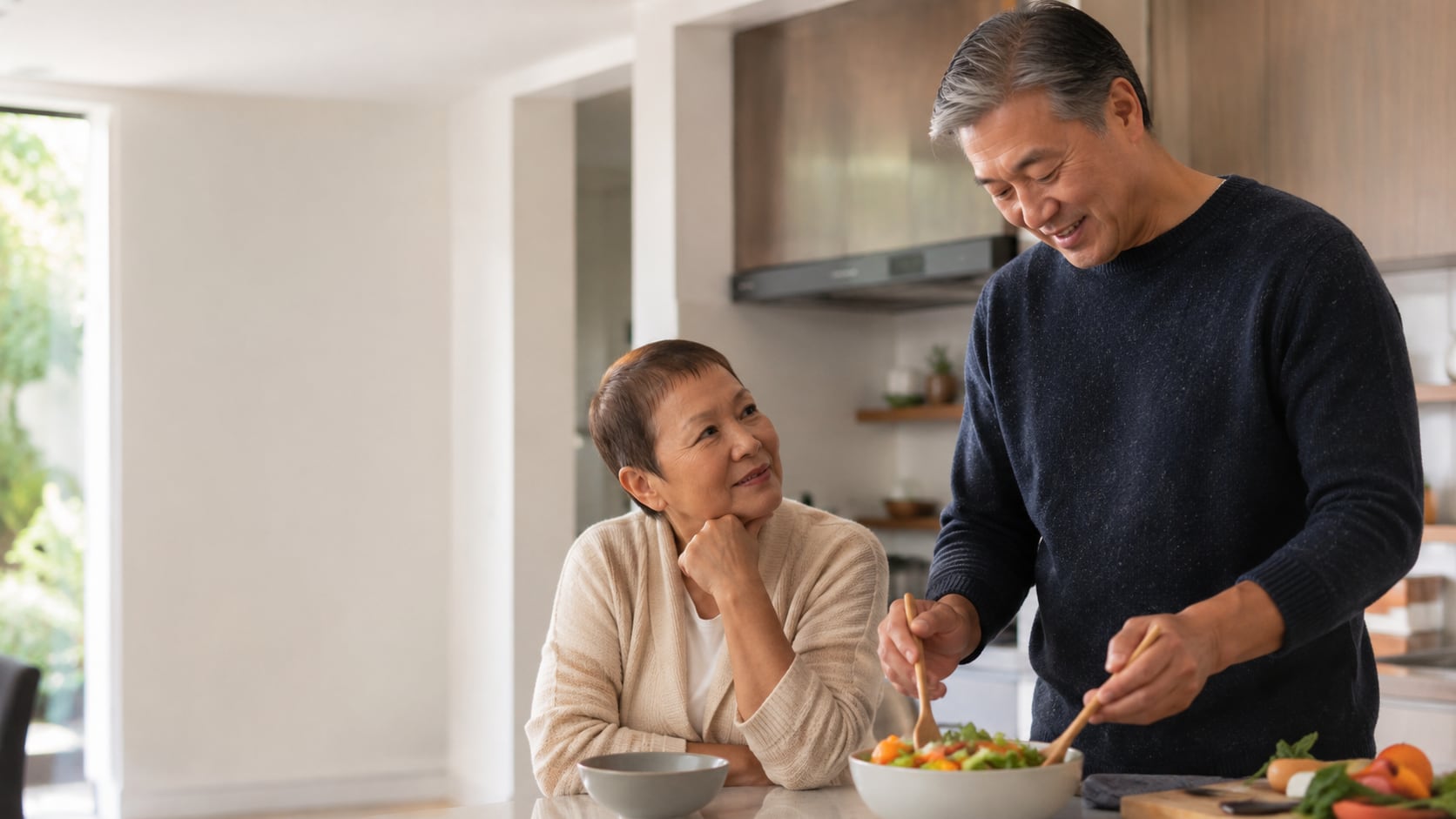 An older man and woman stand together in a kitchen, the man stirring a bowl of salad while the woman smiles up at him.