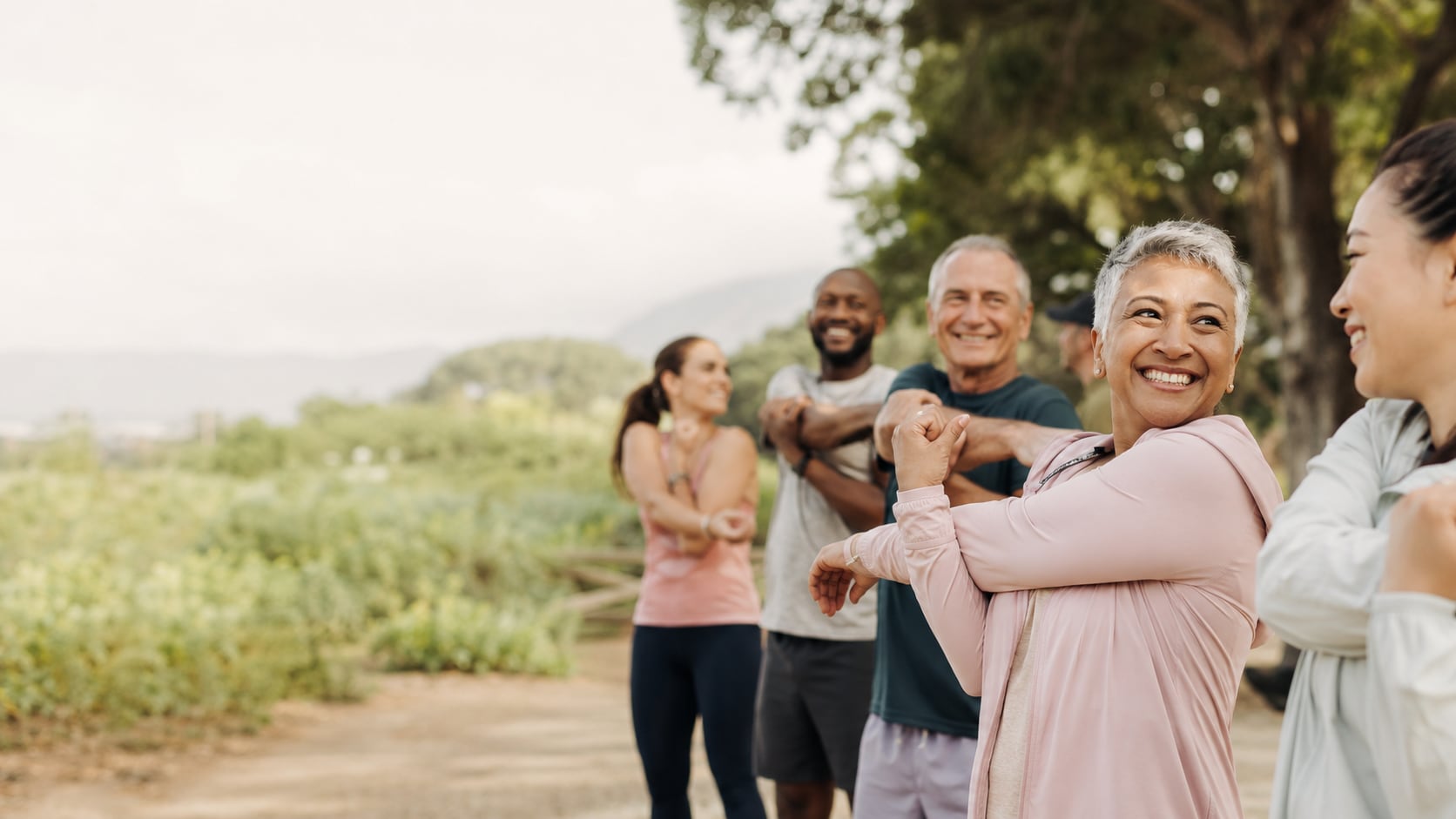 A group of adults of mixed ages stretching together outdoors on a grassy path, laughing.