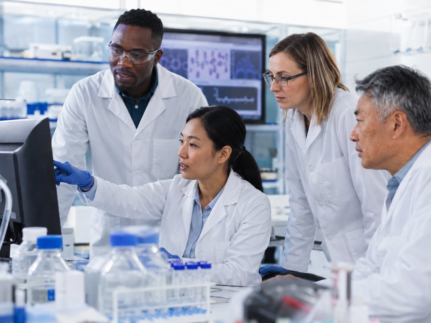 Four lab researchers in coats examine data on a computer monitor together in a laboratory.