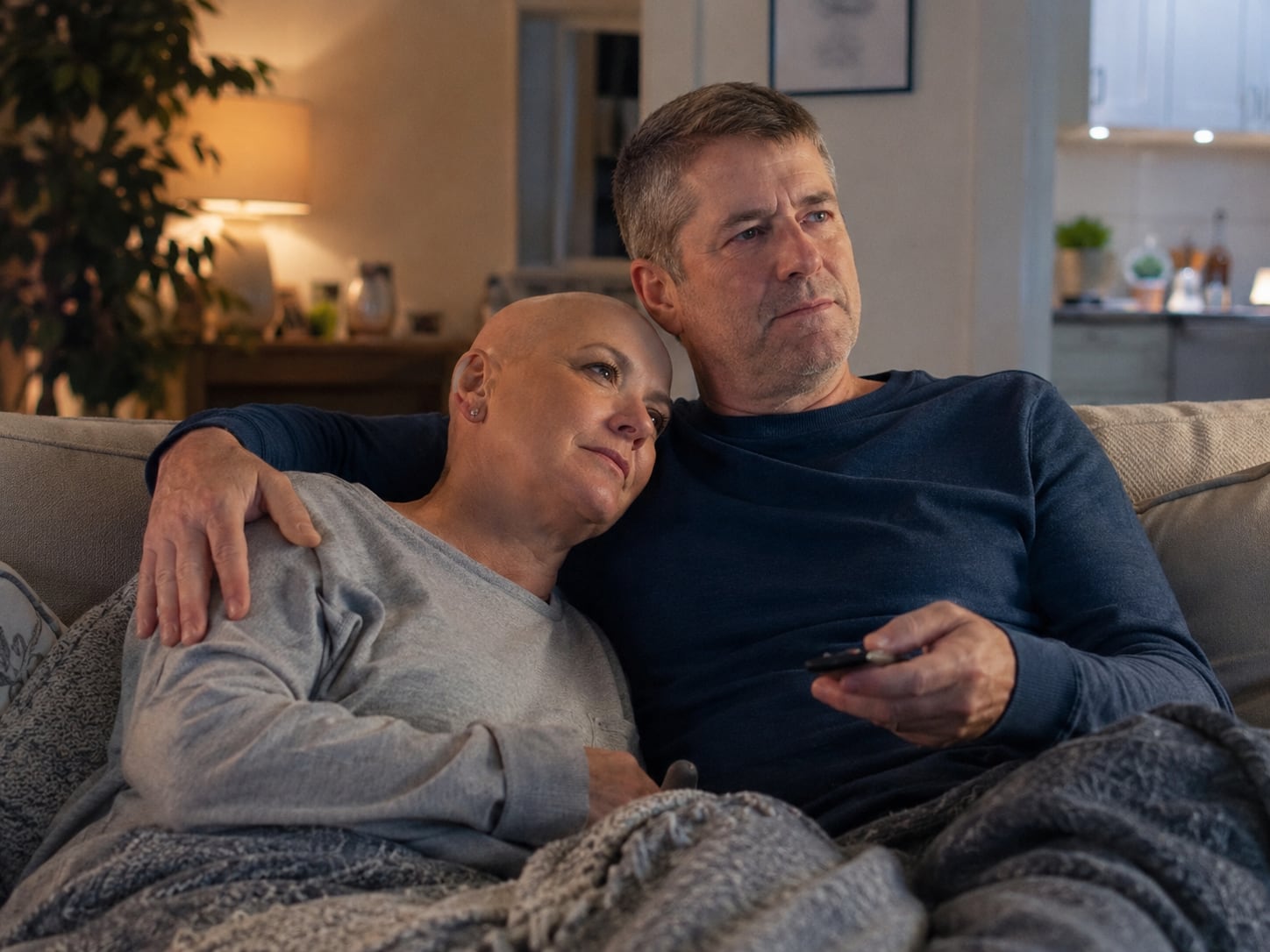 A woman with a shaved head rests her head on a man's shoulder as they sit together on a couch.