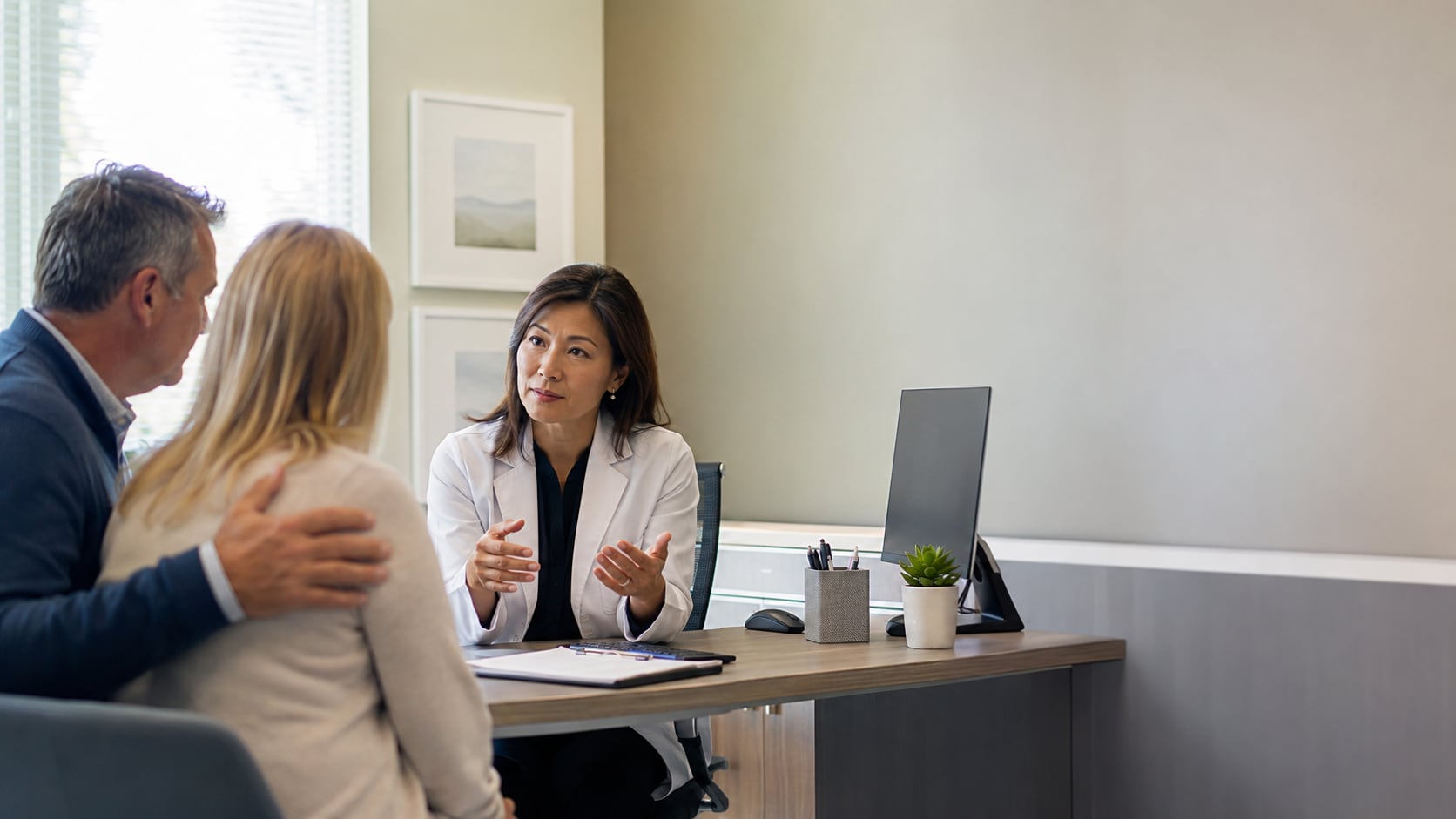 A female doctor in a white coat sits at a desk gesturing while talking with a man and woman, her hand resting on the woman's shoulder.