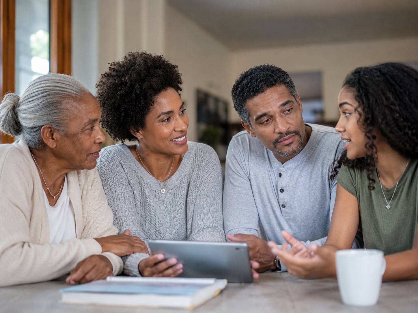 Four people sit together at a table looking at a tablet, in conversation at home.
