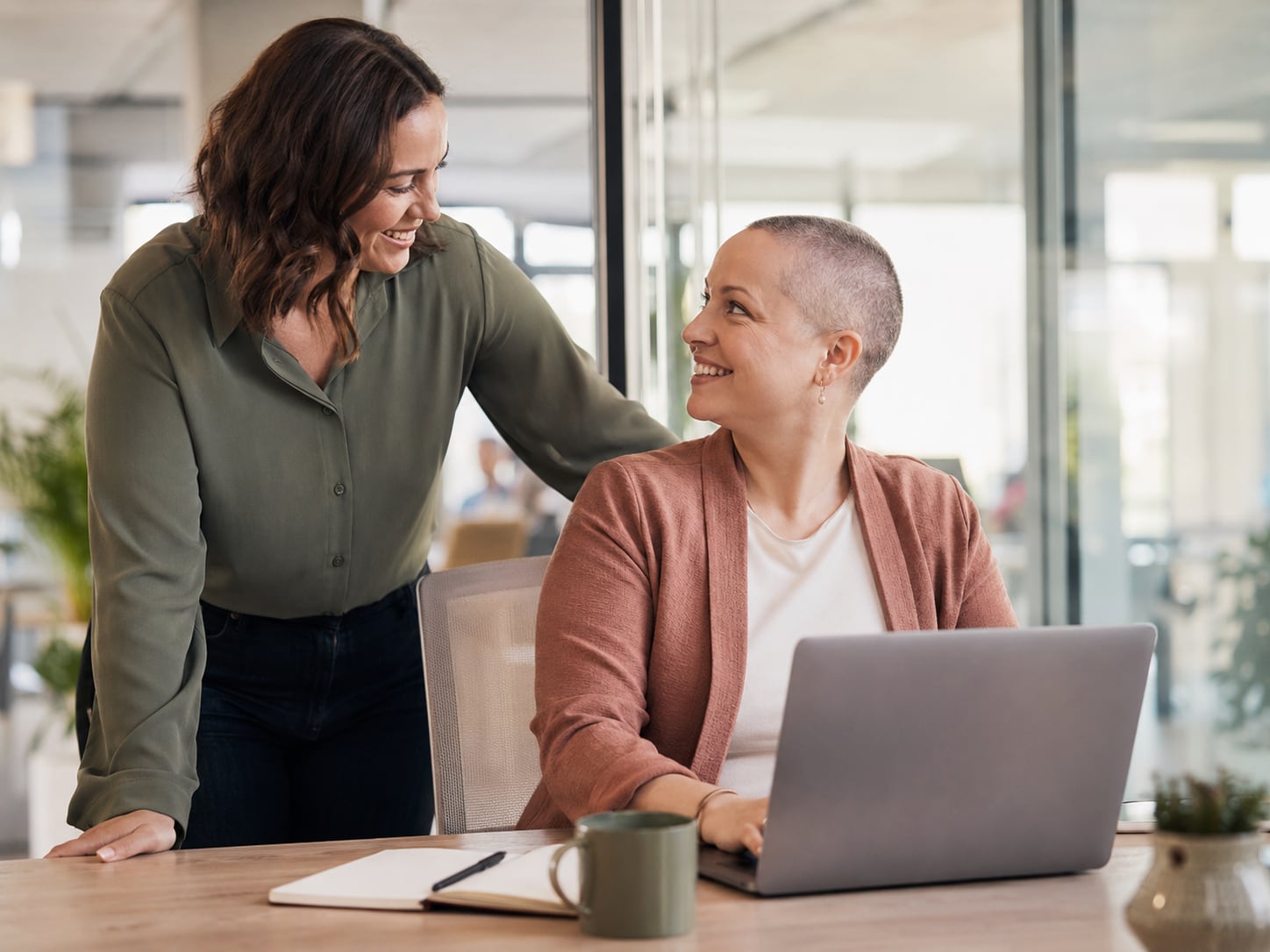 A woman with a shaved head sits at a laptop while a colleague stands behind her, hand on her shoulder, both smiling.