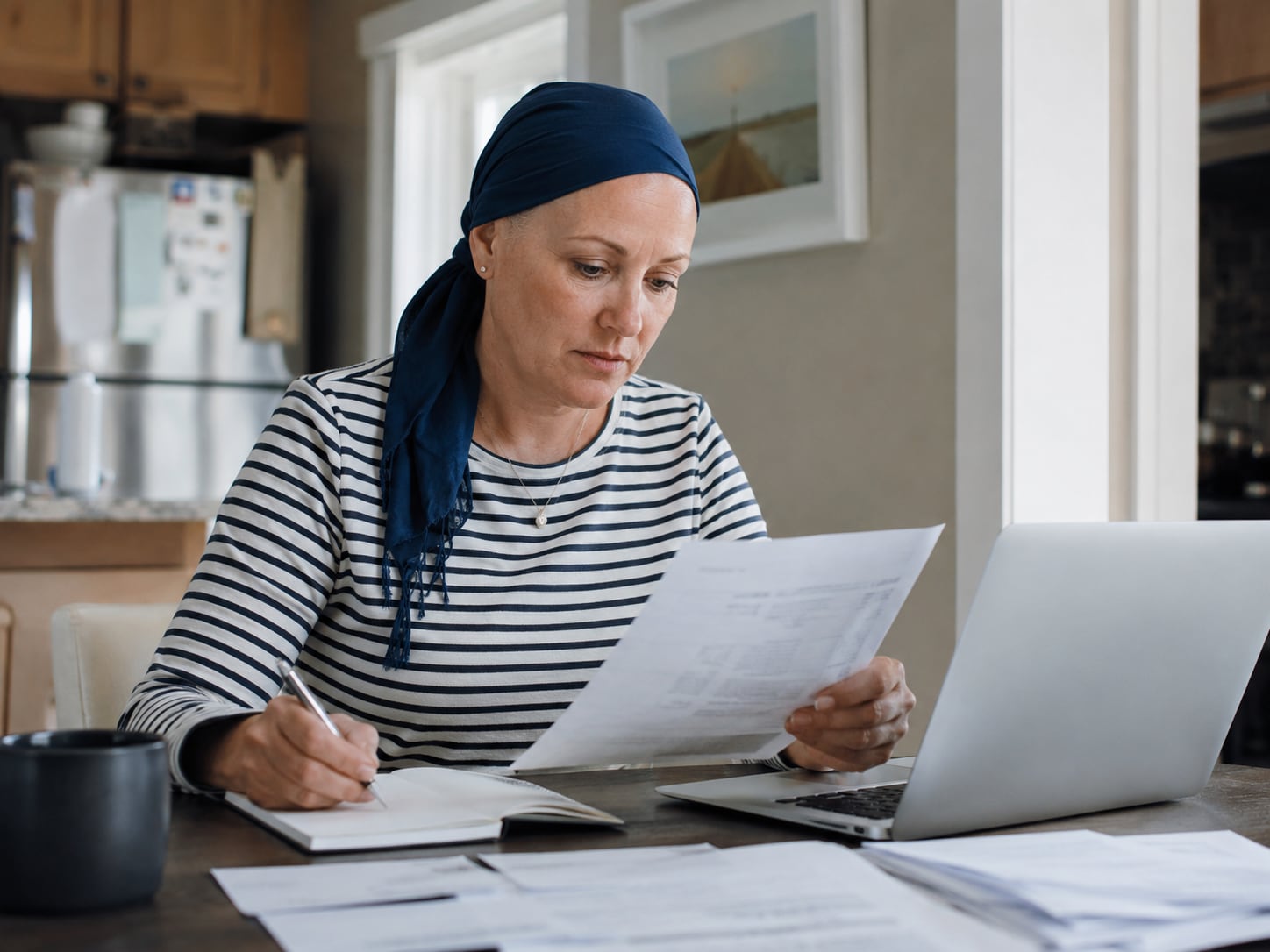 A woman wearing a headscarf sits at a table writing in a notebook, with a document and laptop nearby.