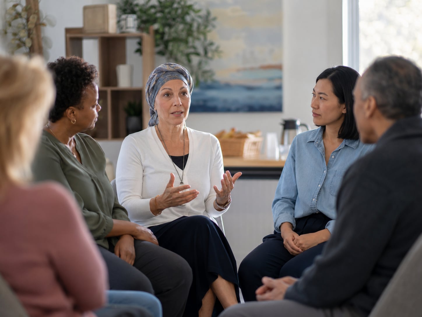 A support group of five people sit together in a living room; a woman wearing a headscarf gestures while speaking, and the others, both women and a man, listen.