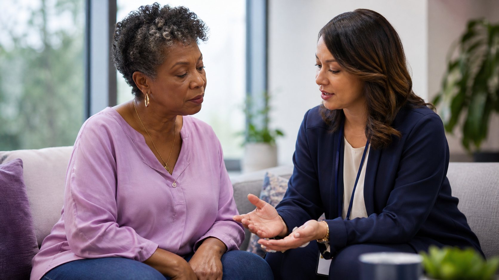 A patient navigator with a lanyard explains a sequence of steps to an older patient seated on a sofa.