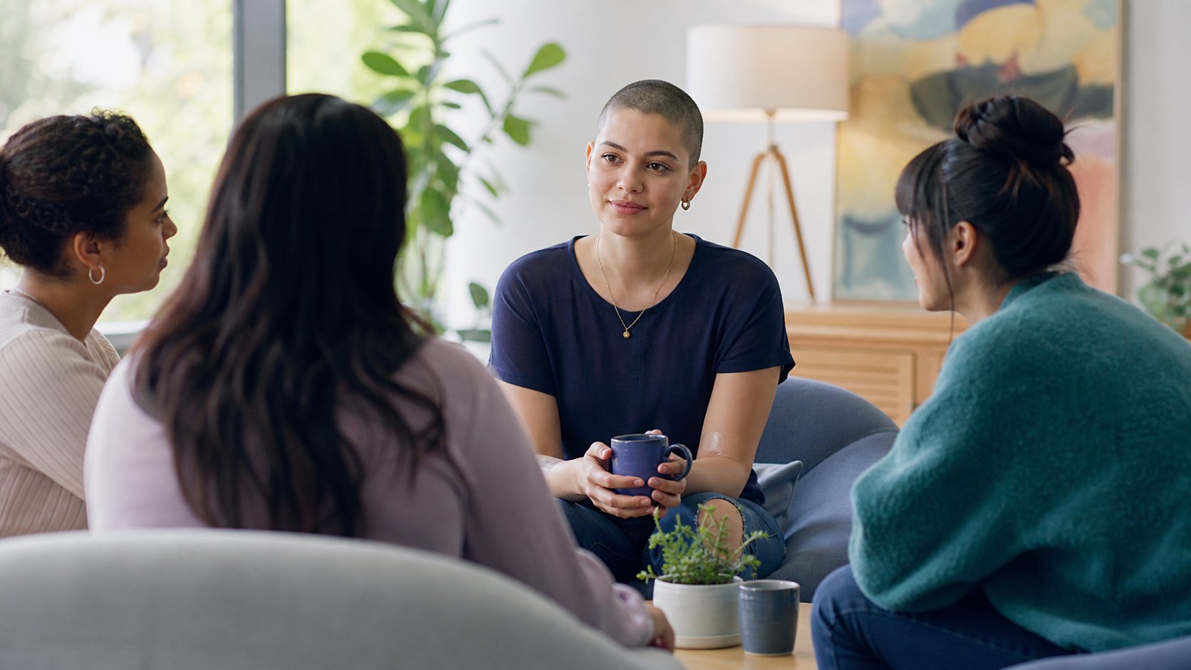 A young adult with a shaved head talks with peers in a bright living room.