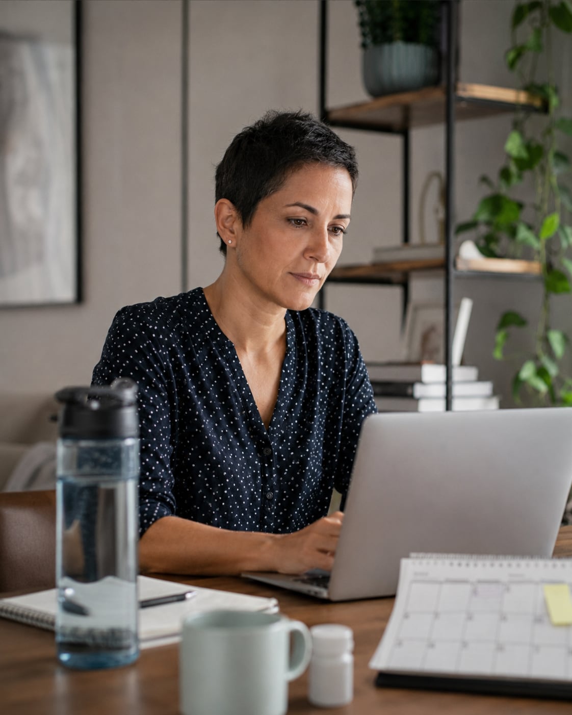 A woman sits at a desk with a laptop, looking down thoughtfully, alone.