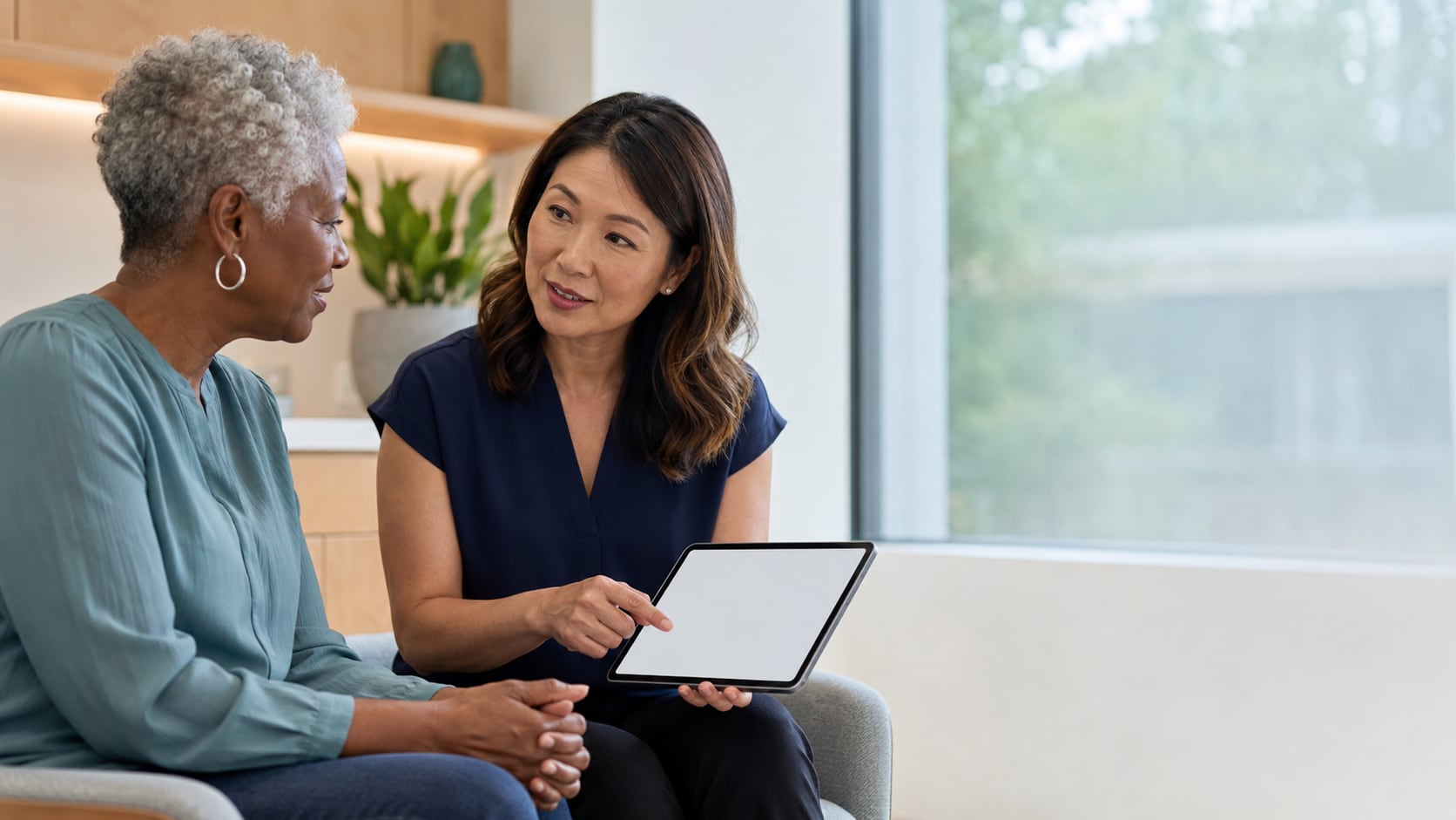 An older woman and a female clinician sit together by a window; the clinician points to information on a tablet as they talk.