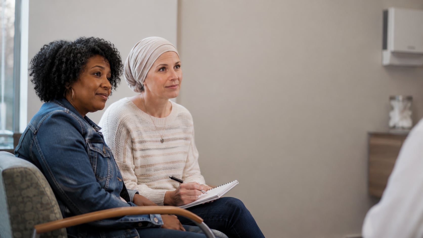 Two women sit side by side in a clinic room, one in a headscarf writing in a notebook, both listening to someone out of frame.