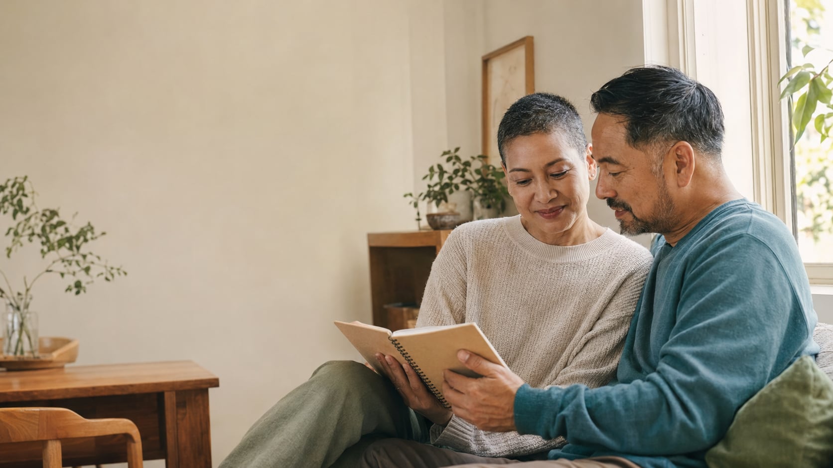 An older couple sit close together on a sofa by a window, reading the same book and smiling.