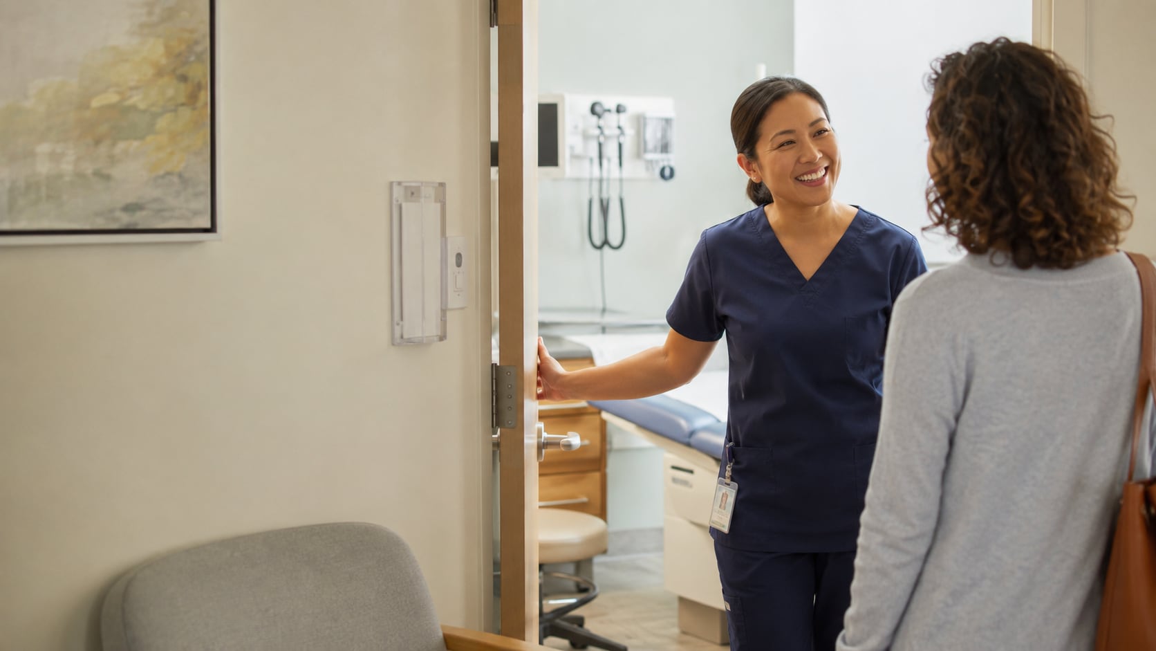 A smiling nurse holds open the door to an exam room, greeting a patient standing in the hallway.