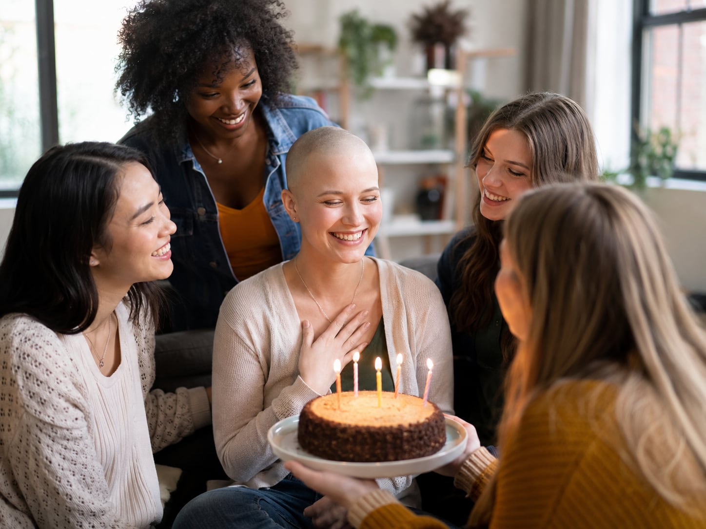 A group of women celebrate with a lit cake around a woman with a shaved head, hand on her chest, smiling.